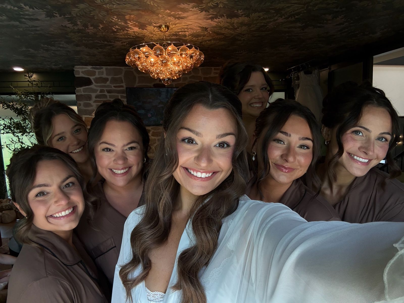 A bride in a white robe surrounded by her six bridesmaids in blush robes on the morning of the wedding — every face done by Samantha