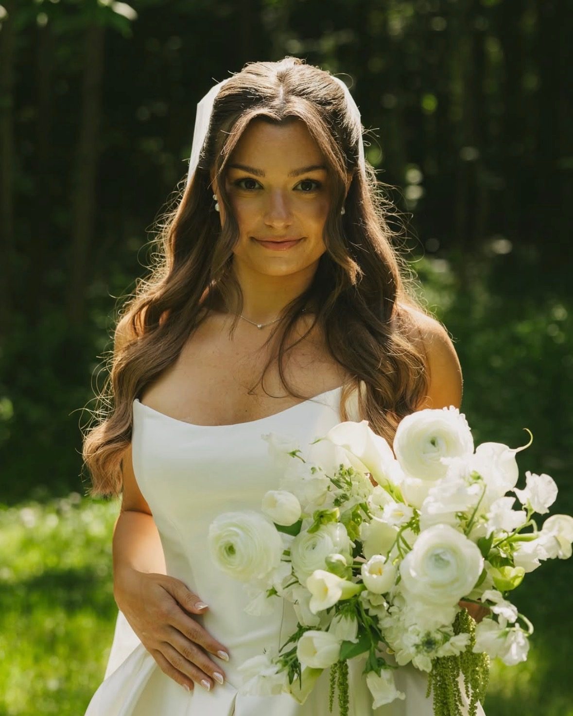 Bride in a soft natural-glam look, half-up hair, holding a white bouquet in front of greenery