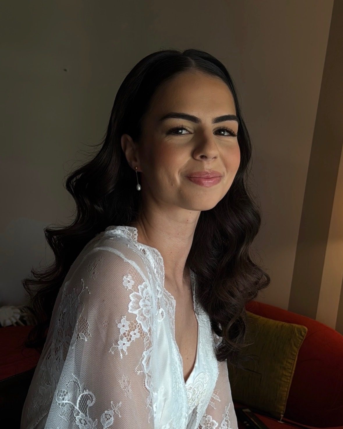 Bride in a lace robe with soft waves and defined glam makeup, warm window light